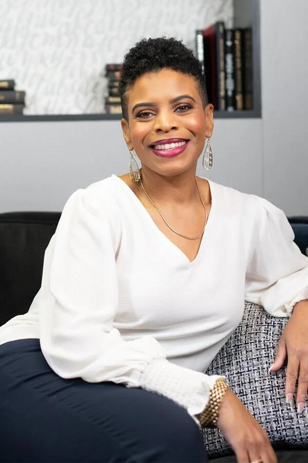 Stacey Mahoney Photo Stacey Mahoney smiling on a sofa in front of a bookshelf. She is wearing a white blouse, navy pants, and dangling gold earrings.